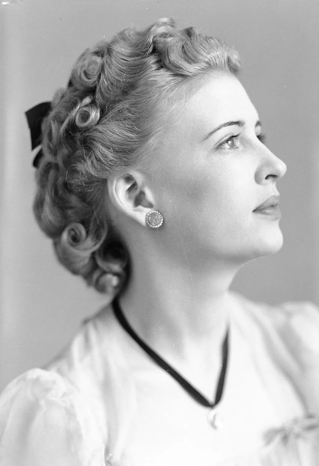 Portrait of a woman with short light curled hair wthi a dark bow, and wears a white blouse with puff sleeves and a necklace with a thick dark band, and large round earrings. She is oriented to her left and looks off and up to her left, with her face almost in profile.