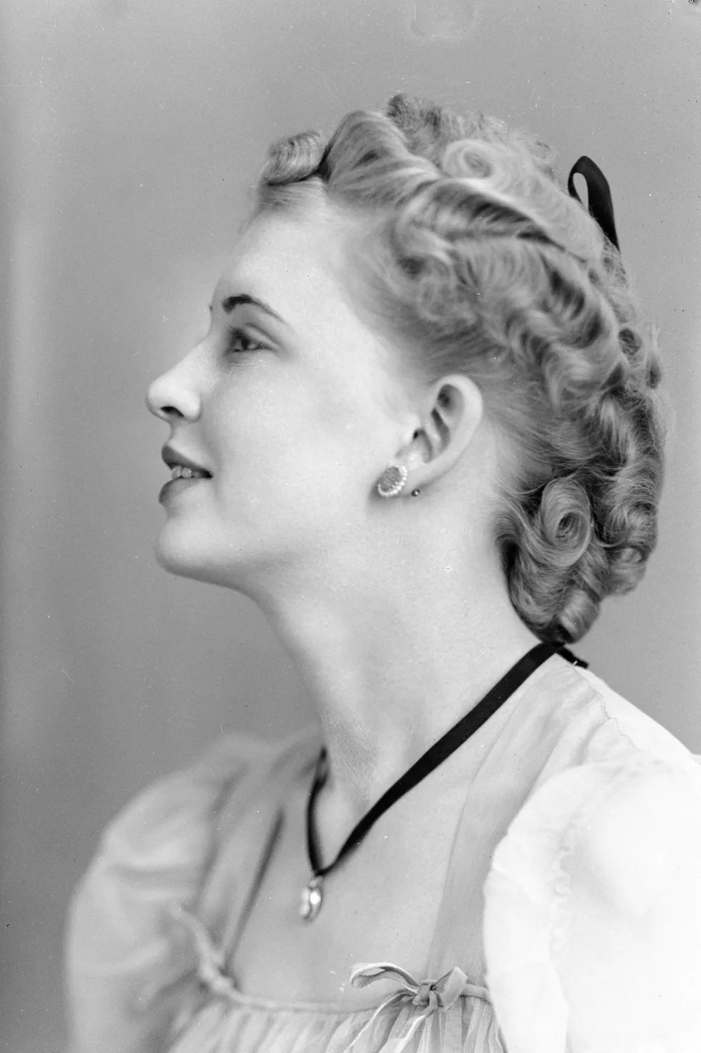 Portrait of a woman with short light curled hair wthi a dark bow, and wears a white blouse with puff sleeves and bows on either side of the square neckline, and a necklace with a thick dark band and large round earrings. She is oriented to her right and looks off and up to her right, with her face in profile.