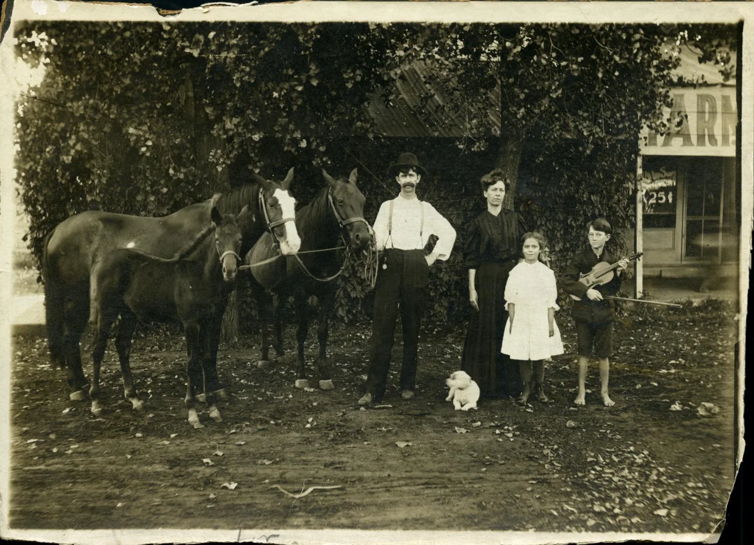 a family of one man one woman and two children standing in from of a store with three horses. The young male child has a violin and there is a white dog laying beside the young female child.